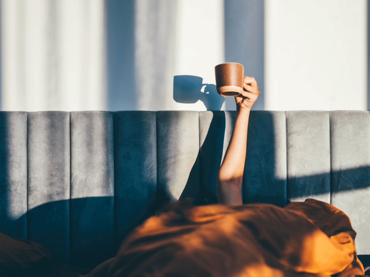 Person sleeping under the covers, holding up a cup of coffee