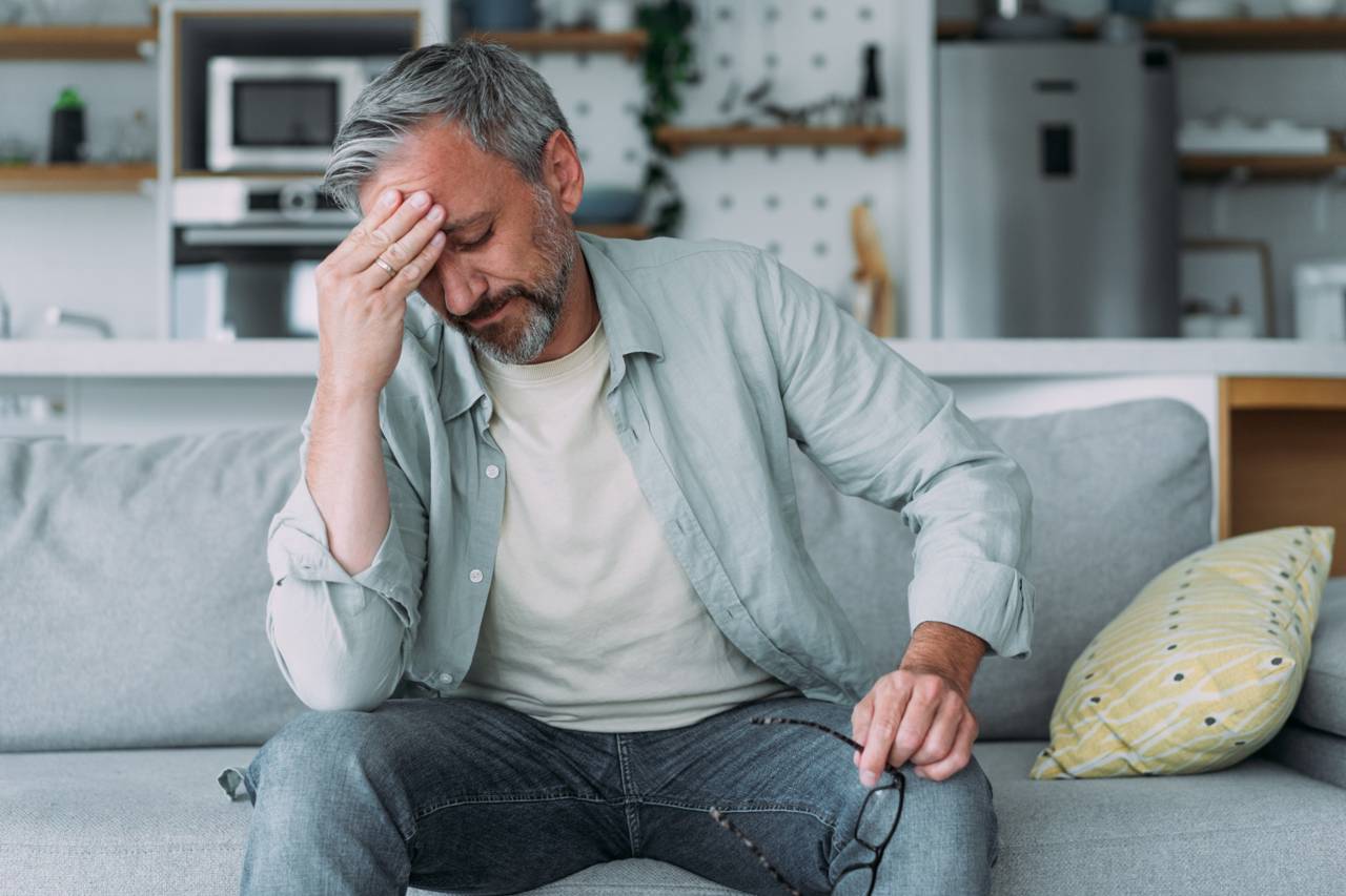 Middle-aged man sitting on a sofa with his hand on his forehead, showing signs of a headache or stress at home.