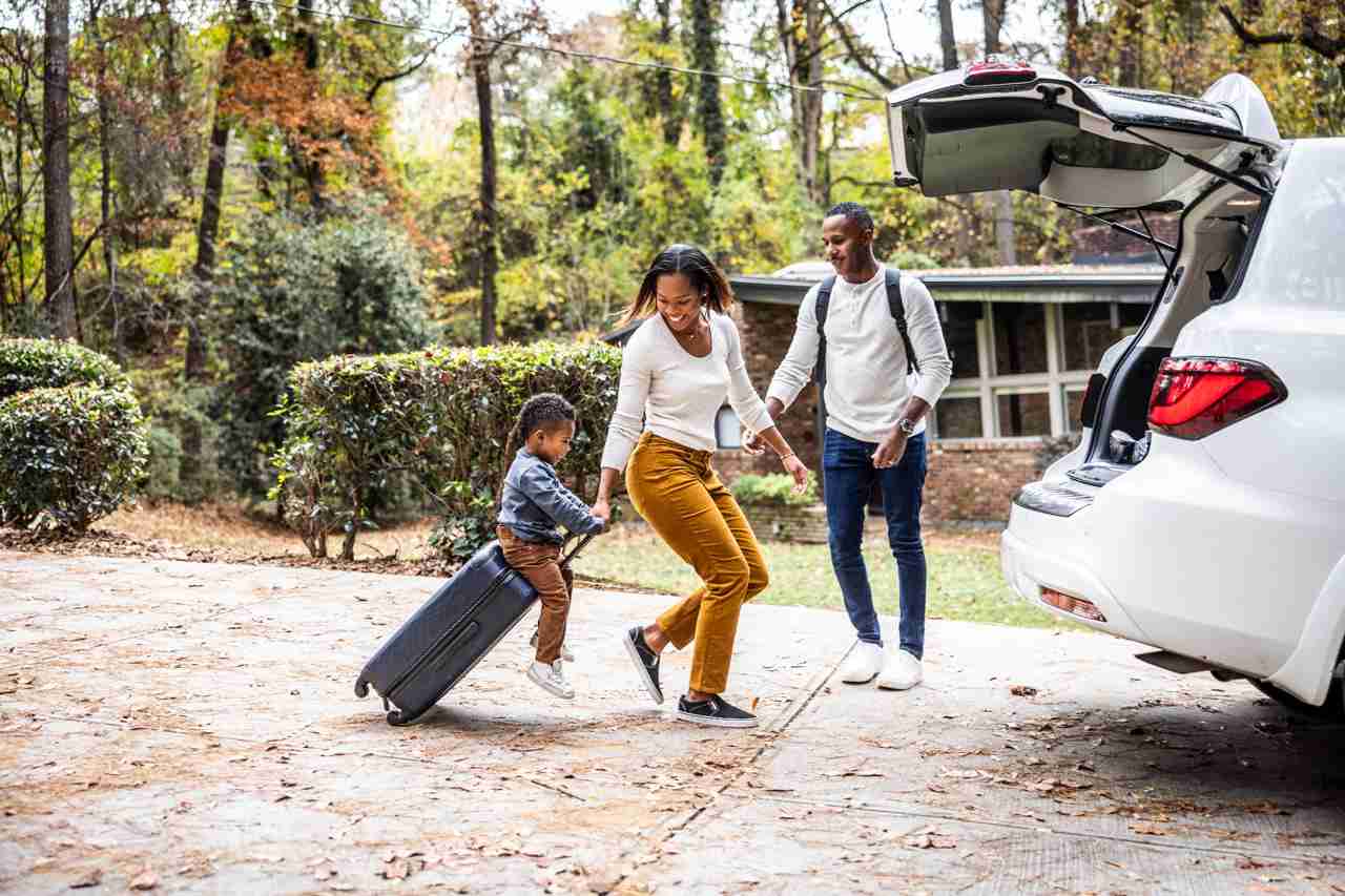 Family with child carrying suitcase while leaving home with car parked