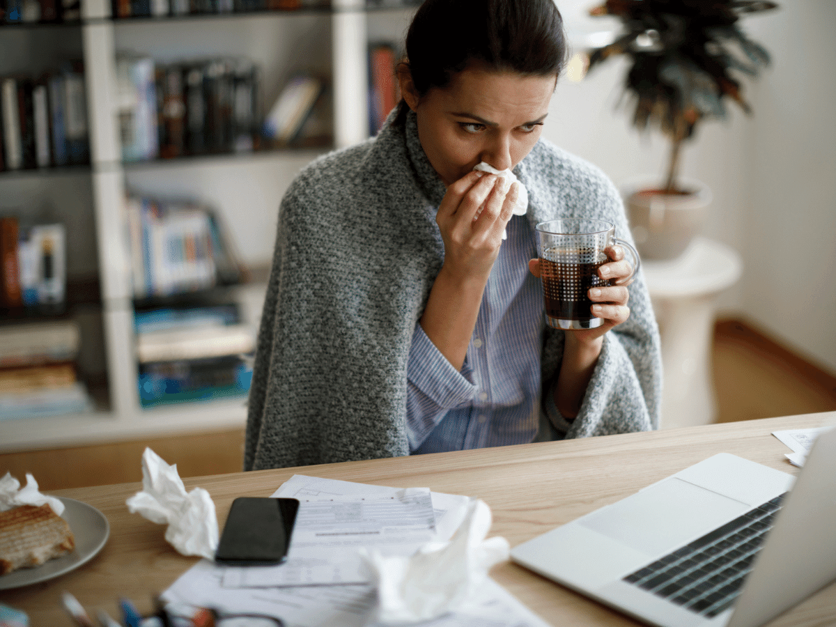 A woman is sick, blowing her nose while sitting at a desk looking at a computer
