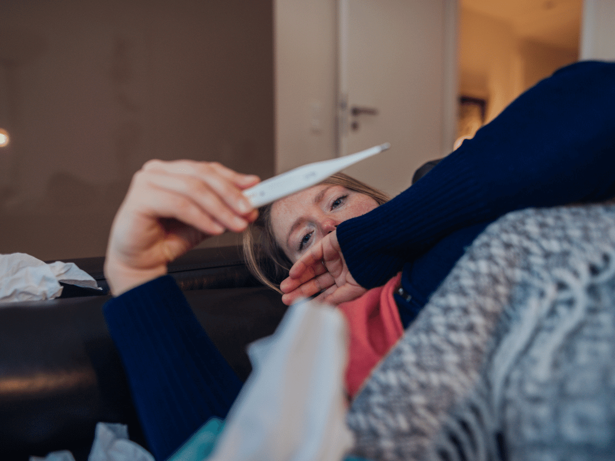 sick woman laying on the couch looking at a thermometer