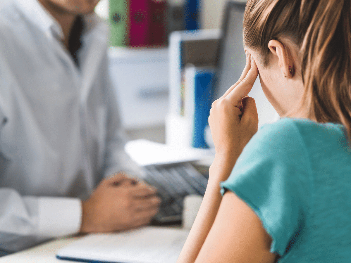 Young woman holds her head in pain while talking with a doctor