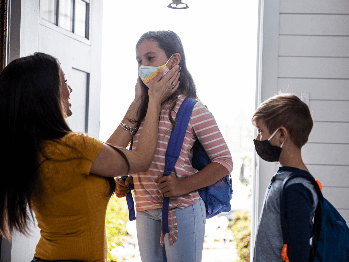 Mother helping two young children get ready for school, putting cloth masks on before leaving the house