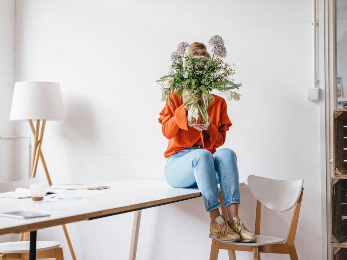 Woman sitting on a table hiding her face behind a vase of flowers
