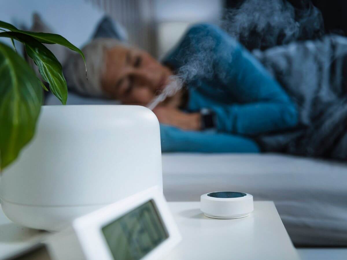 In the foreground, a humidifier on a night stand is running. Out of focus in the background, an older woman is sleeping in bed.
