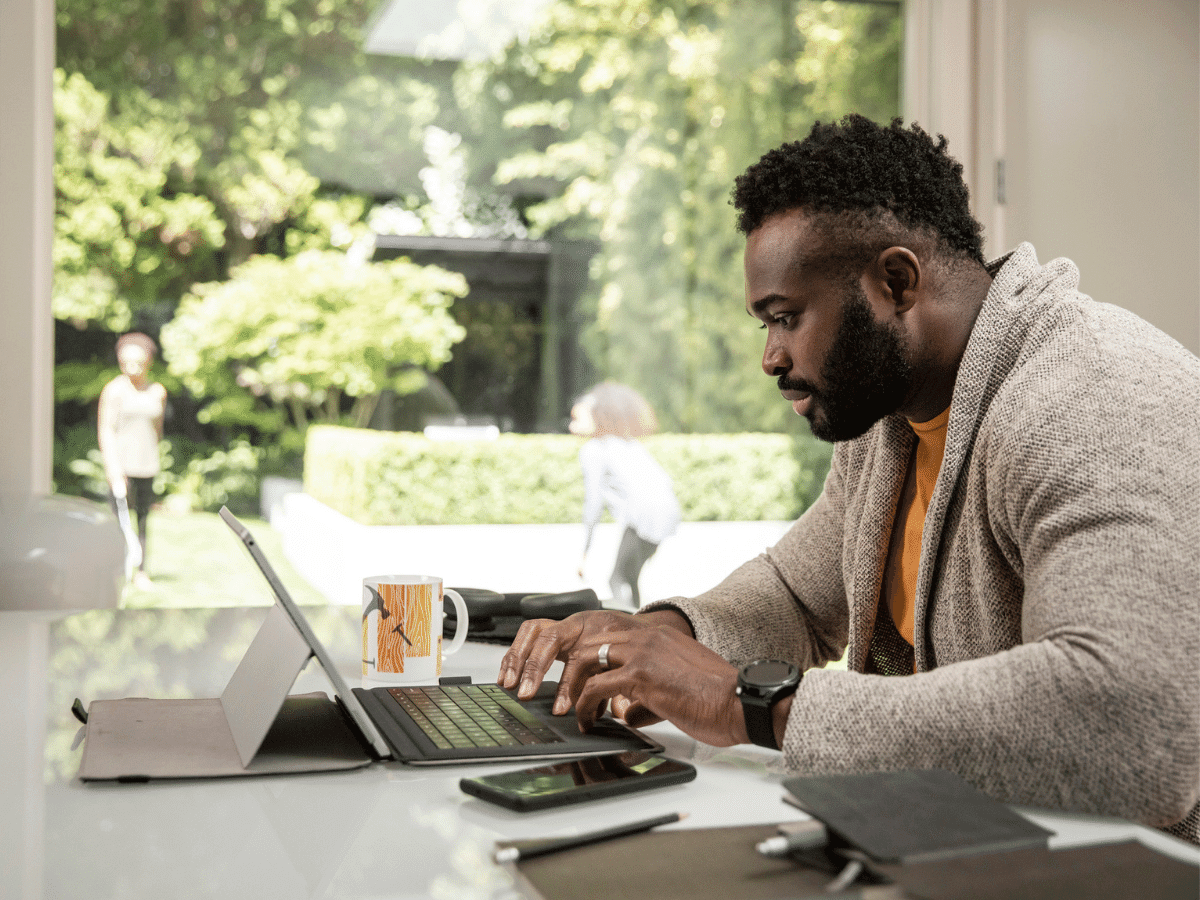 A man sits in his home office typing on his tablet keyboard while his family plays outside in the yard.