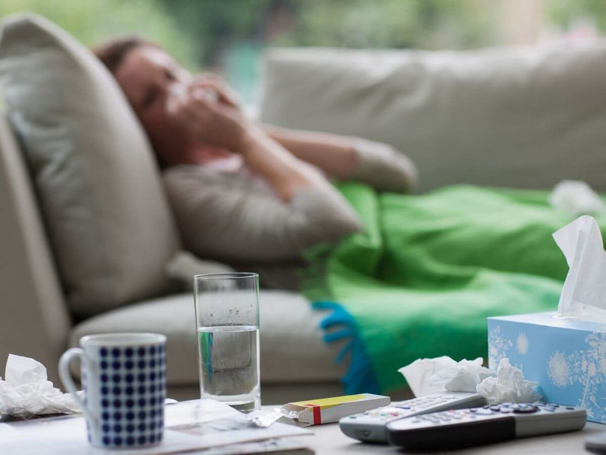 Sick woman lays on the couch in the background of the photo. In the foreground are a box of facial tissues, a cup of water and a box of medication.