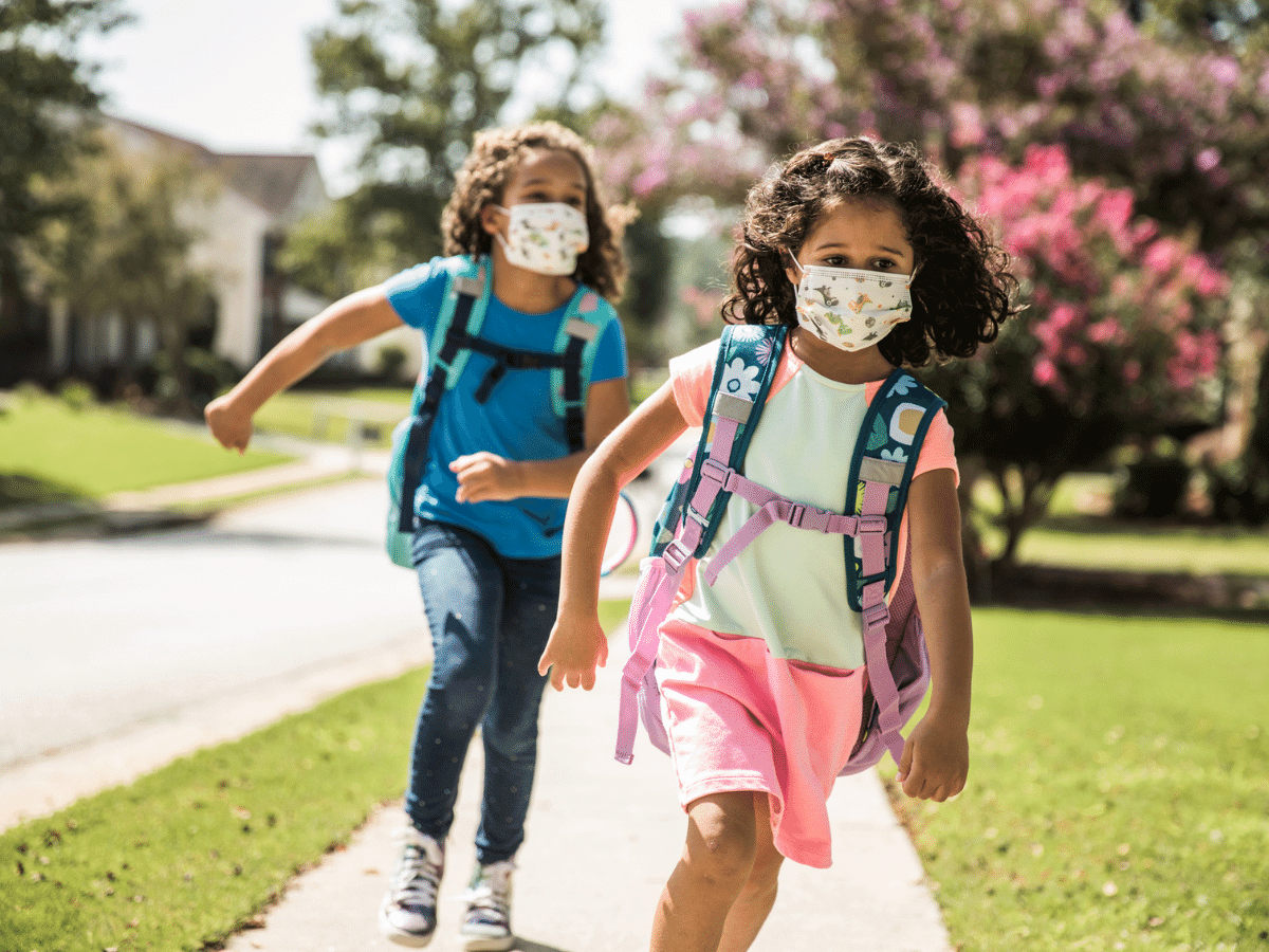 Young girls wearing protective masks and backpacks running on sidewalk