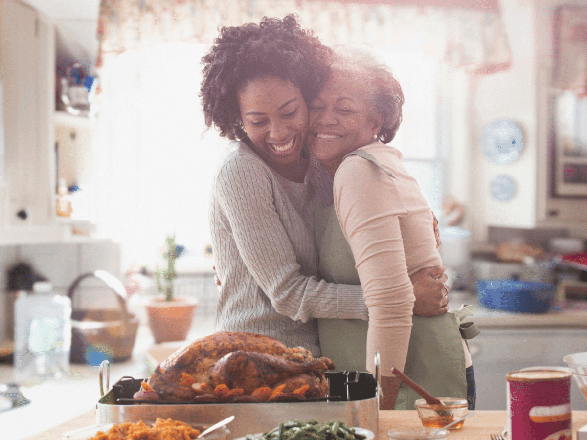 Adult daughter and senior mother hug in the kitchen in front of cooked Thanksgiving turkey