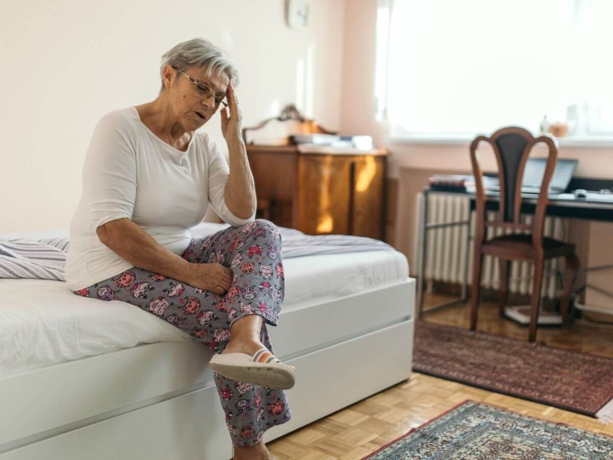 Middle aged woman sitting on the edge of a bed, holding her head in her hand with her eyes closed