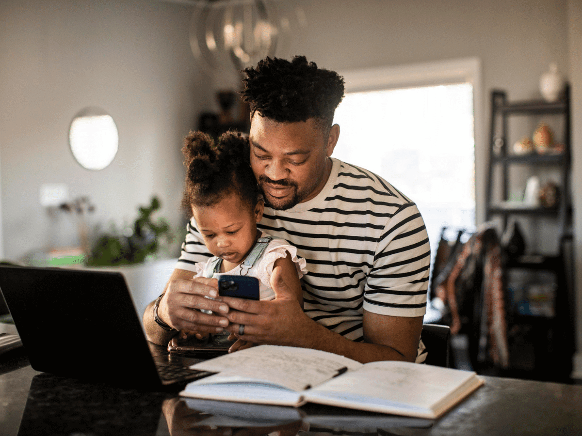 Father holding toddler in his lap with computer open in front of him while he makes a phone call