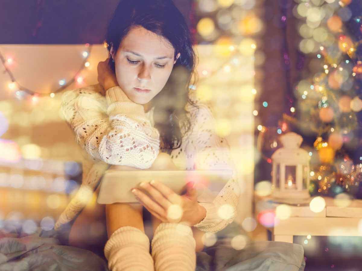 Young woman who looks sad, scrolls on a tablet in front of holiday lights
