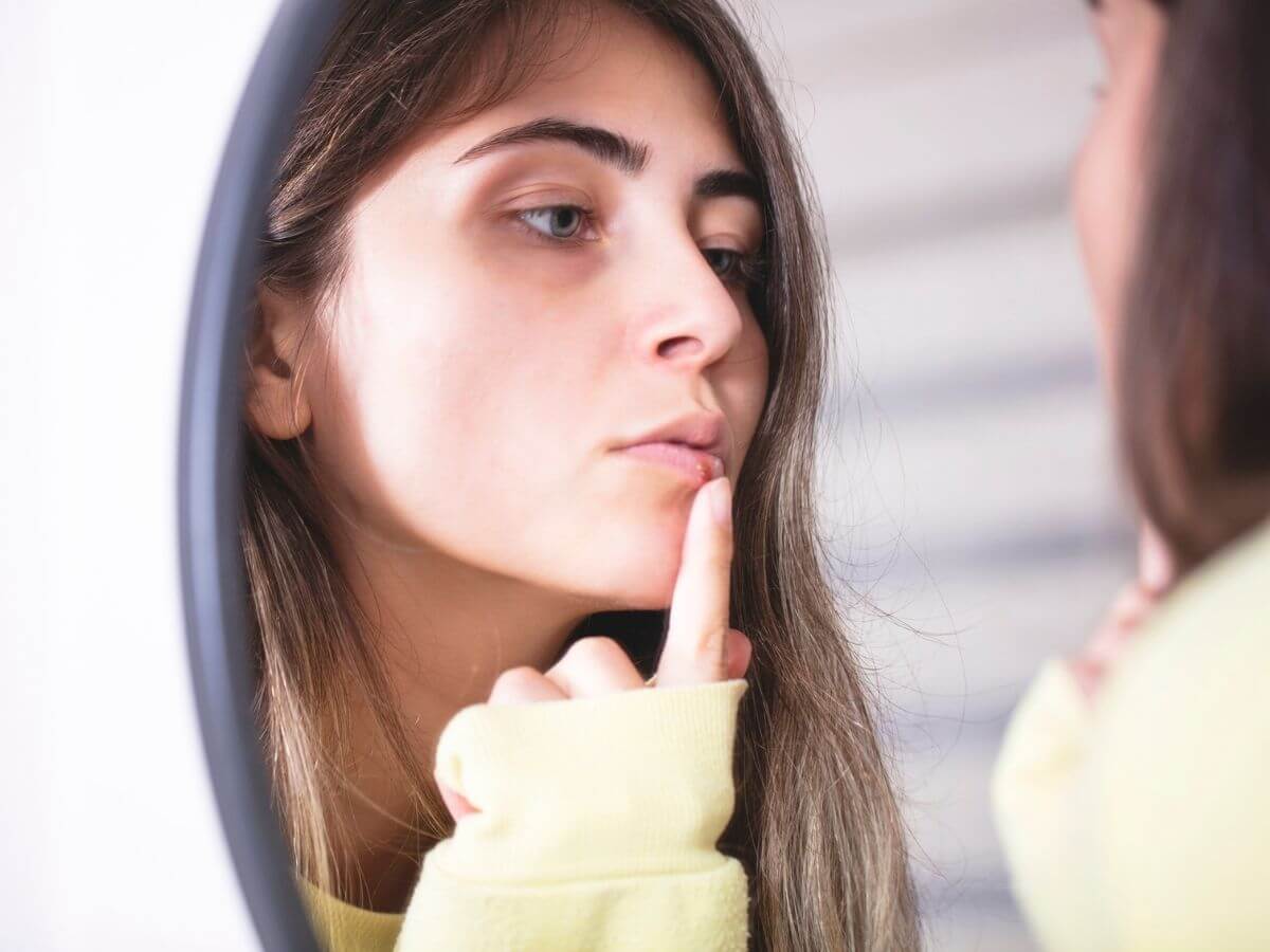 Close up of a young woman looking at her reflection in the mirror and touching an emerging cold sore