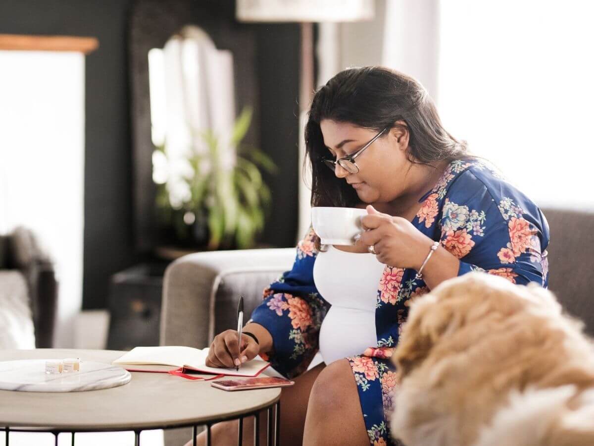 Candid shot of a woman sitting on a couch drinking coffee while writing in a journal. A golden retriever is visible in the foreground.