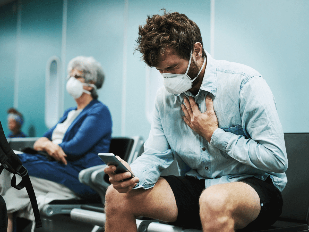 Young man wearing a mask holds his chest in pain while looking at a smartphone in a waiting room.