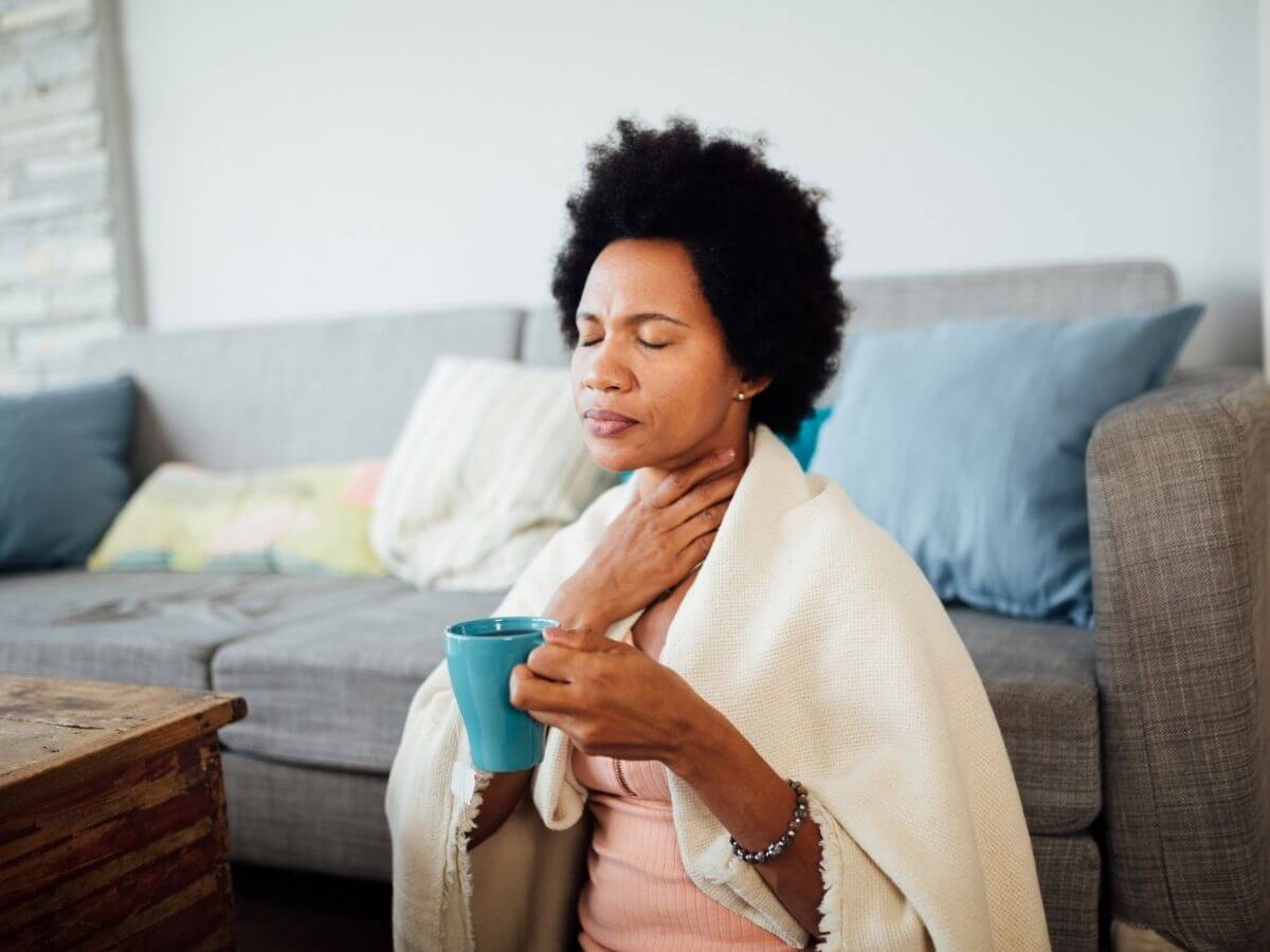 Woman sitting with a blanket around her shoulders in a living room. She is holding a mug of tea with one hand and holding her throat in pain with the other.