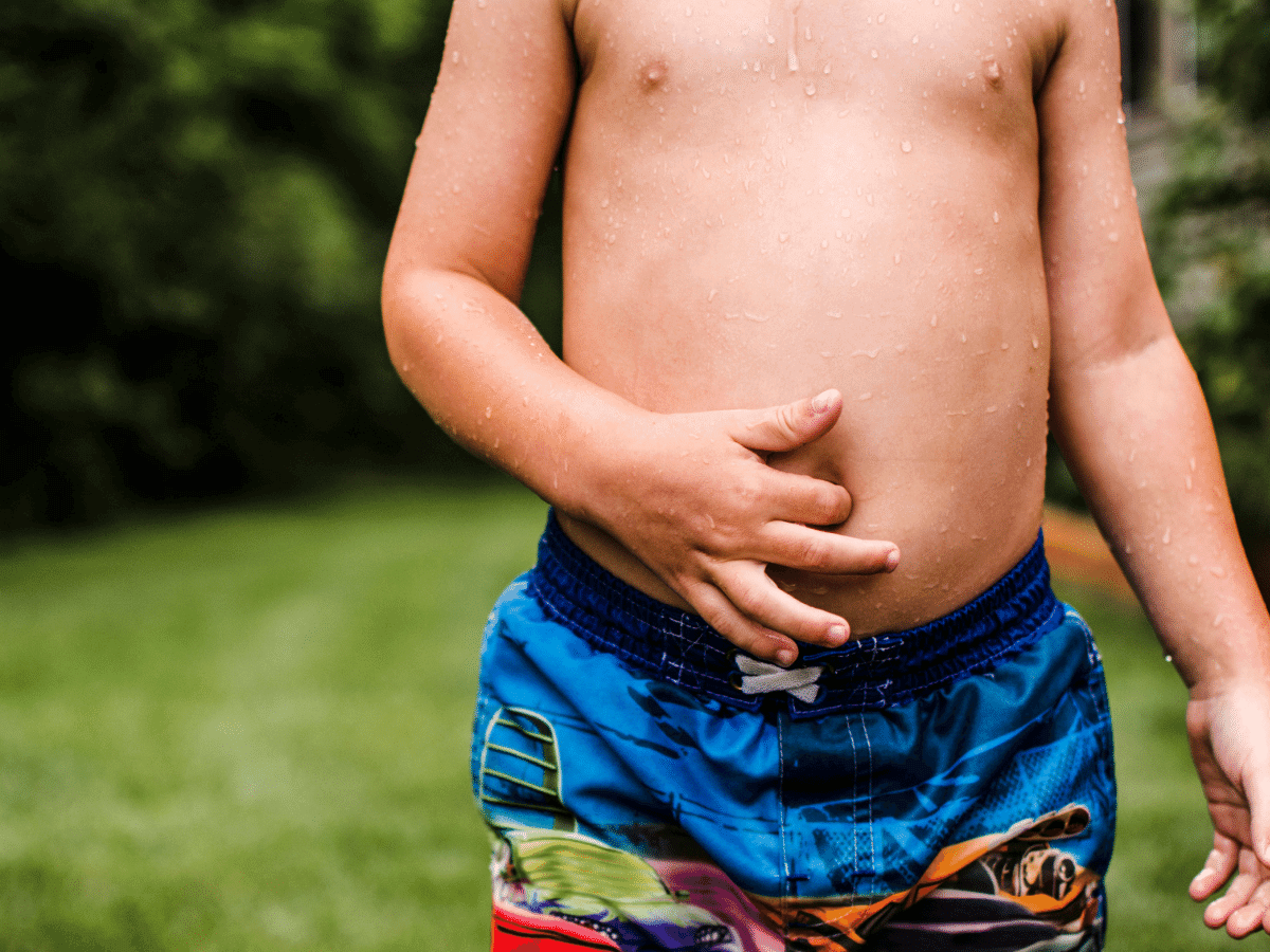 Boy in swim trunks touching his belly button.