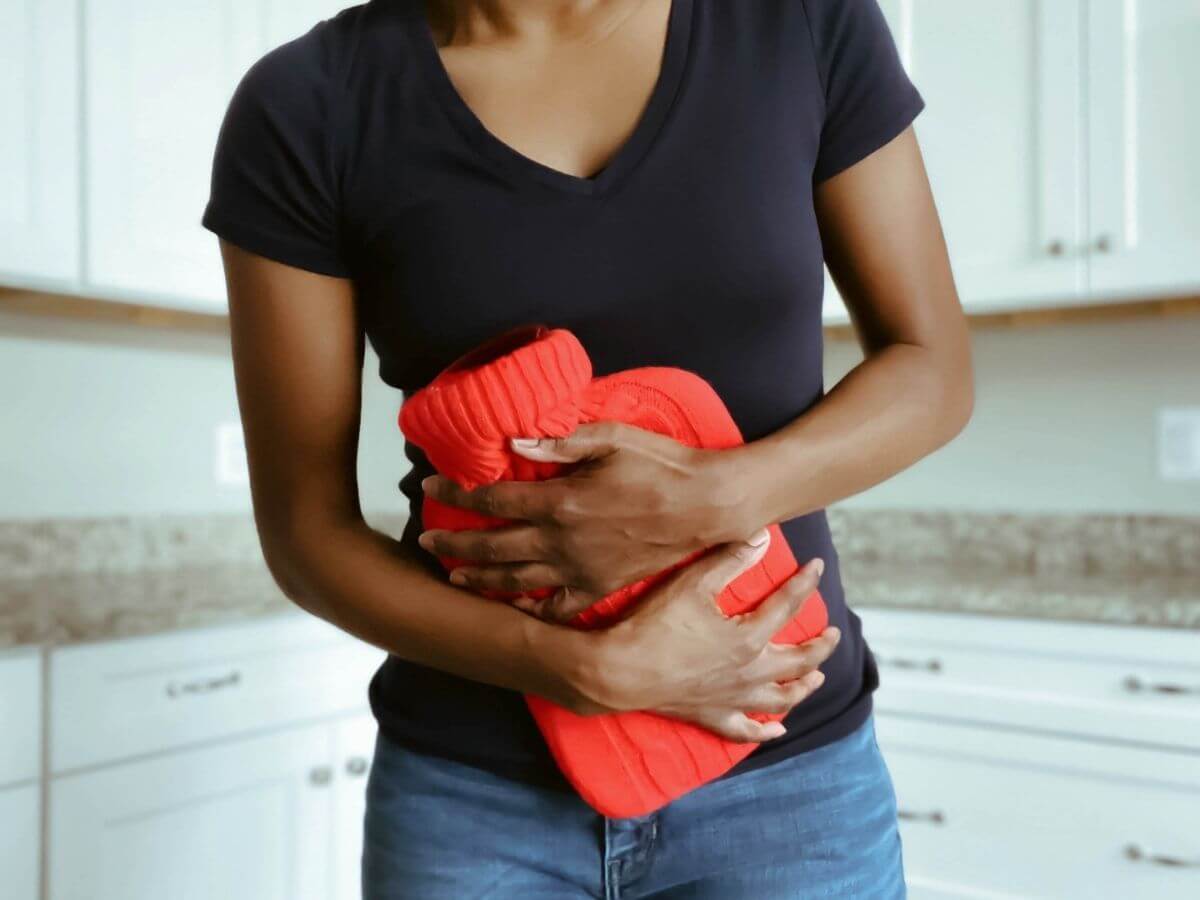 Photo of a woman from the neck down. The woman is holding a hot water bottle on her stomach and appears to be in pain.