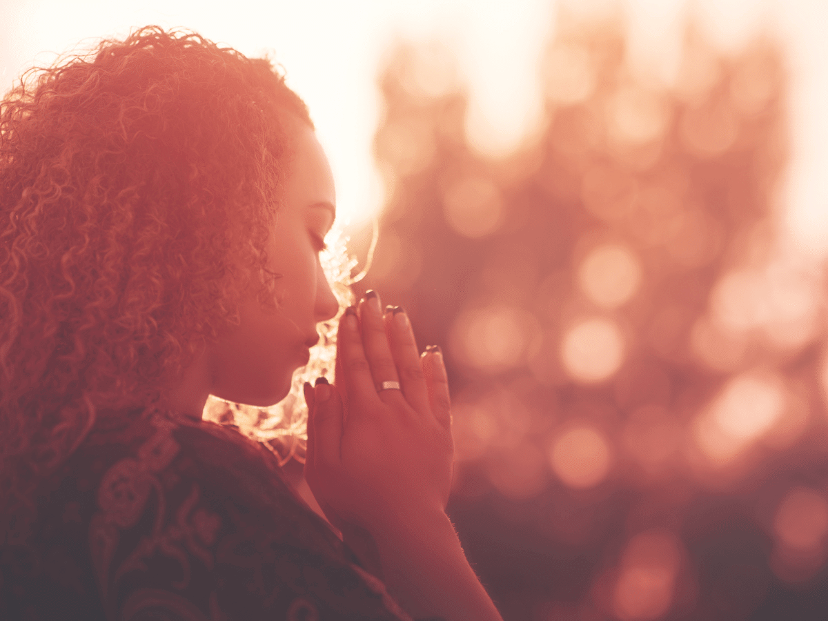Woman praying with her hands clasped and eyes closed at sunset