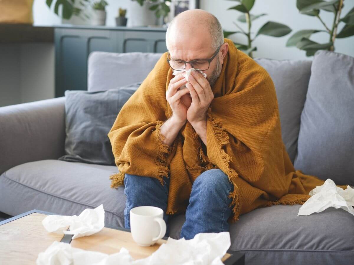middle-aged man sitting on the couch under a blanket. He is surrounded by tissues and is blowing his nose
