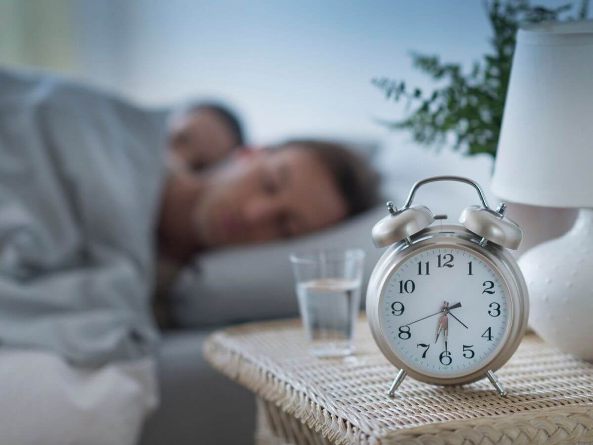 Man and woman sleeping in bed, focus is on the bedside table and alarm clock in the foreground