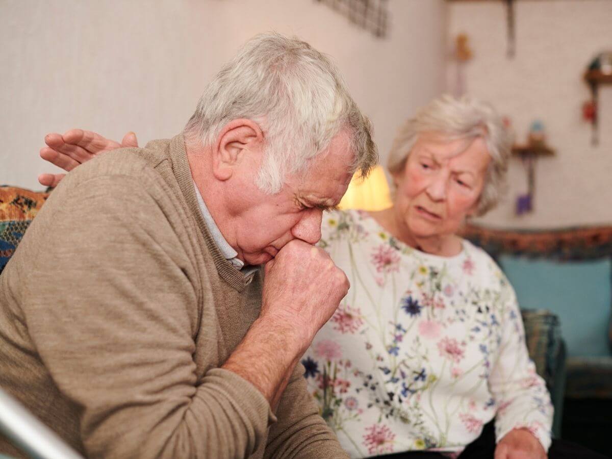Older couple sitting on the couch. The woman comforts the man as he coughs.
