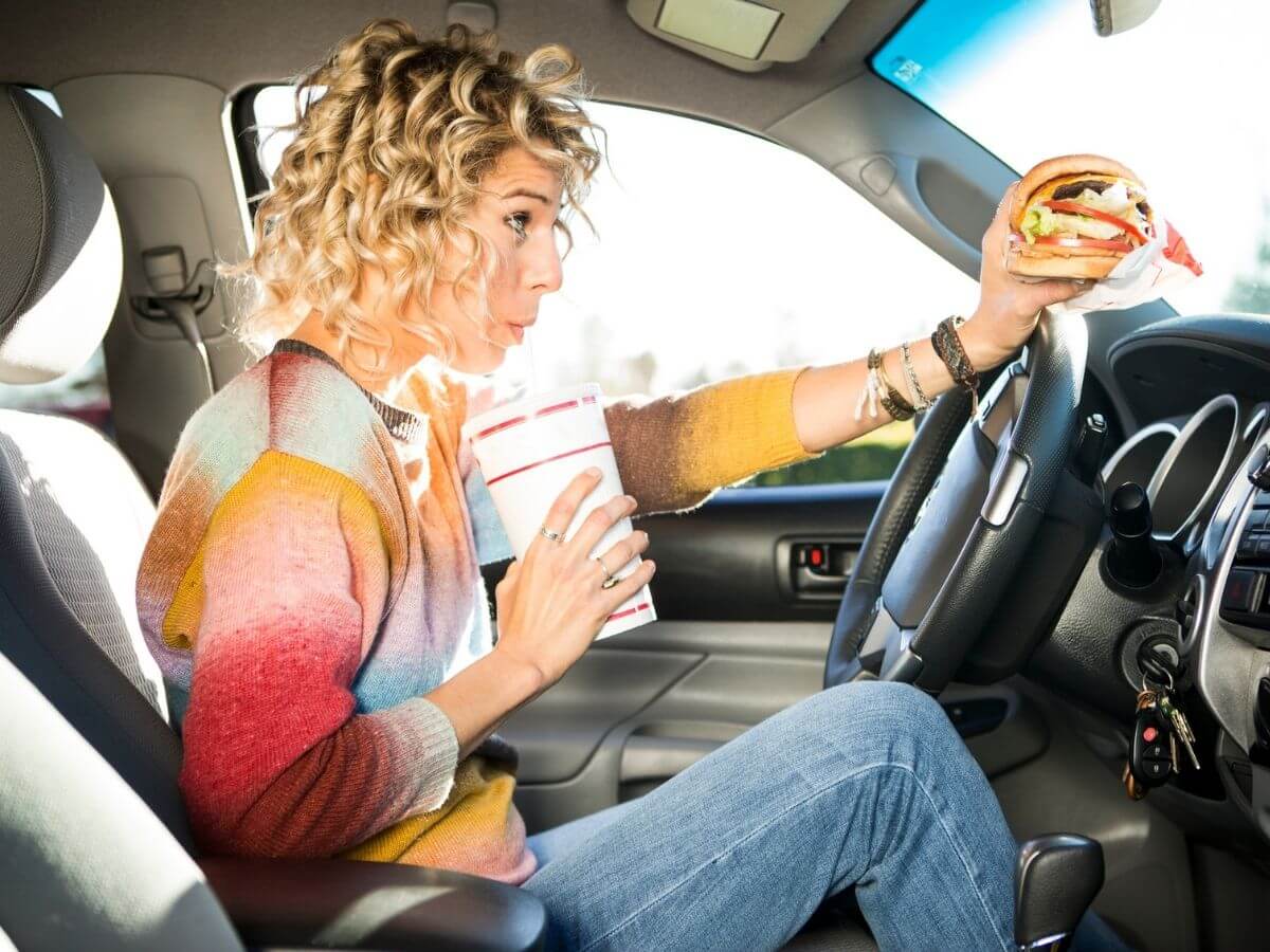 Young woman eating a fast food cheeseburger and soda while driving in her car.