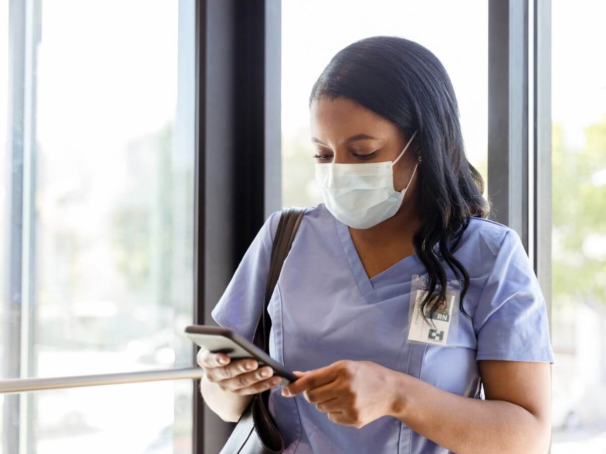 A nurse wearing scrubs and a face mask reads news on her phone in the hallways of a hospital
